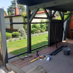 Piotr - Montaż balustrady na tarasie: widoczne narzędzia, poziomica, wkrętarka, wiadro, elementy balustrady, kostka brukowa i trawnik w tle.