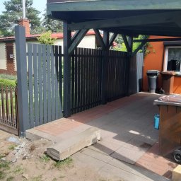 Piotr - Nowoczesne ogrodzenie panelowe w kolorze grafitowym z bramą wjazdową, widoczne z zewnątrz posesji, z elementami kostki brukowej i betonowymi obrzeżami, fragment domu z elewacją w tle.