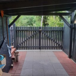 Piotr - Zadaszona wiata z drewnianą bramą pomalowaną na ciemnoszaro, widoczne narzędzia ogrodnicze i materiały budowlane, podłoże wyłożone szarą kostką brukową z ceglanym obramowaniem.