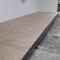 Piotr - Wyłożony jasnymi płytkami taras przed budynkiem, widoczne detale wykończenia krawędzi i otoczenie w trakcie prac budowlanych.