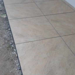 Piotr - Ułożona podłoga z jasnych, kwadratowych płytek ceramicznych z ciemnymi fugami, widoczne niedokończone krawędzie i surowy grunt.