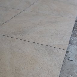 Piotr - Beżowe płytki podłogowe o fakturze kamienia, ułożone w regularny wzór, widoczne szczeliny między płytkami, krawędź z surowym betonem.