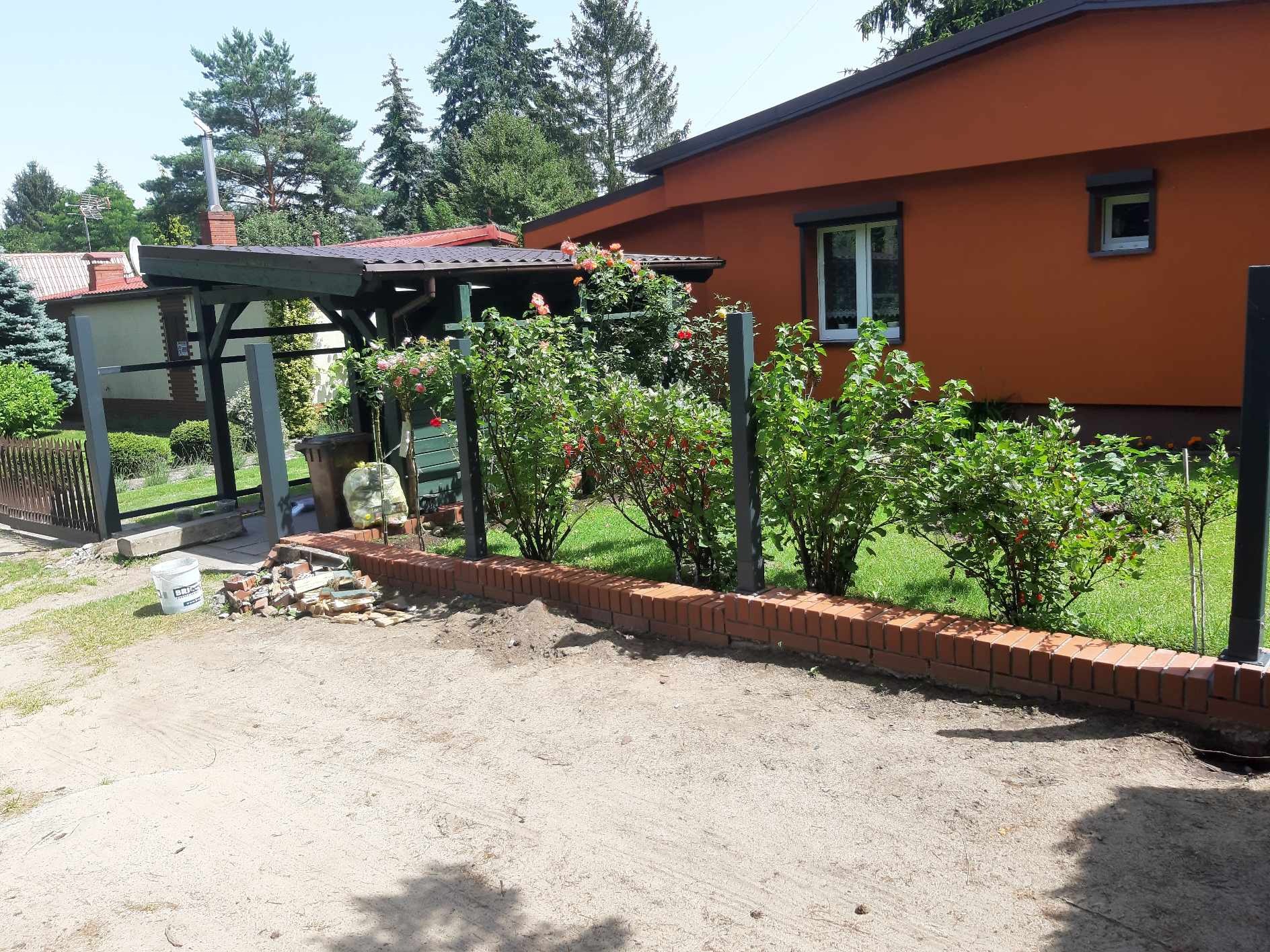 Niski murek z czerwonej cegły oddzielający posesję od drogi, z widocznymi pozostałościami po budowie i pomalowanym na pomarańczowo domem w tle.
