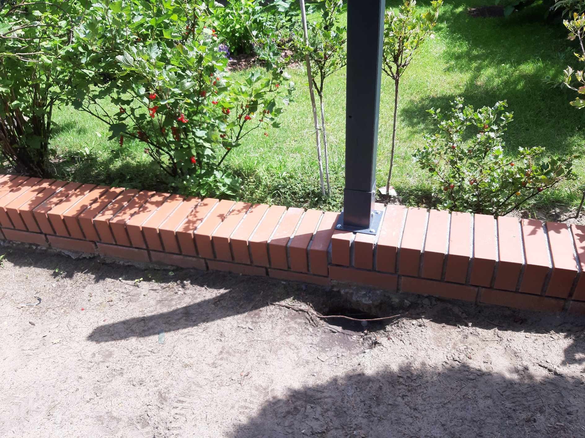 Niski mur z cegły klinkierowej, obok metalowy słupek, widoczne prace ziemne i nasadzenia roślinności w tle.