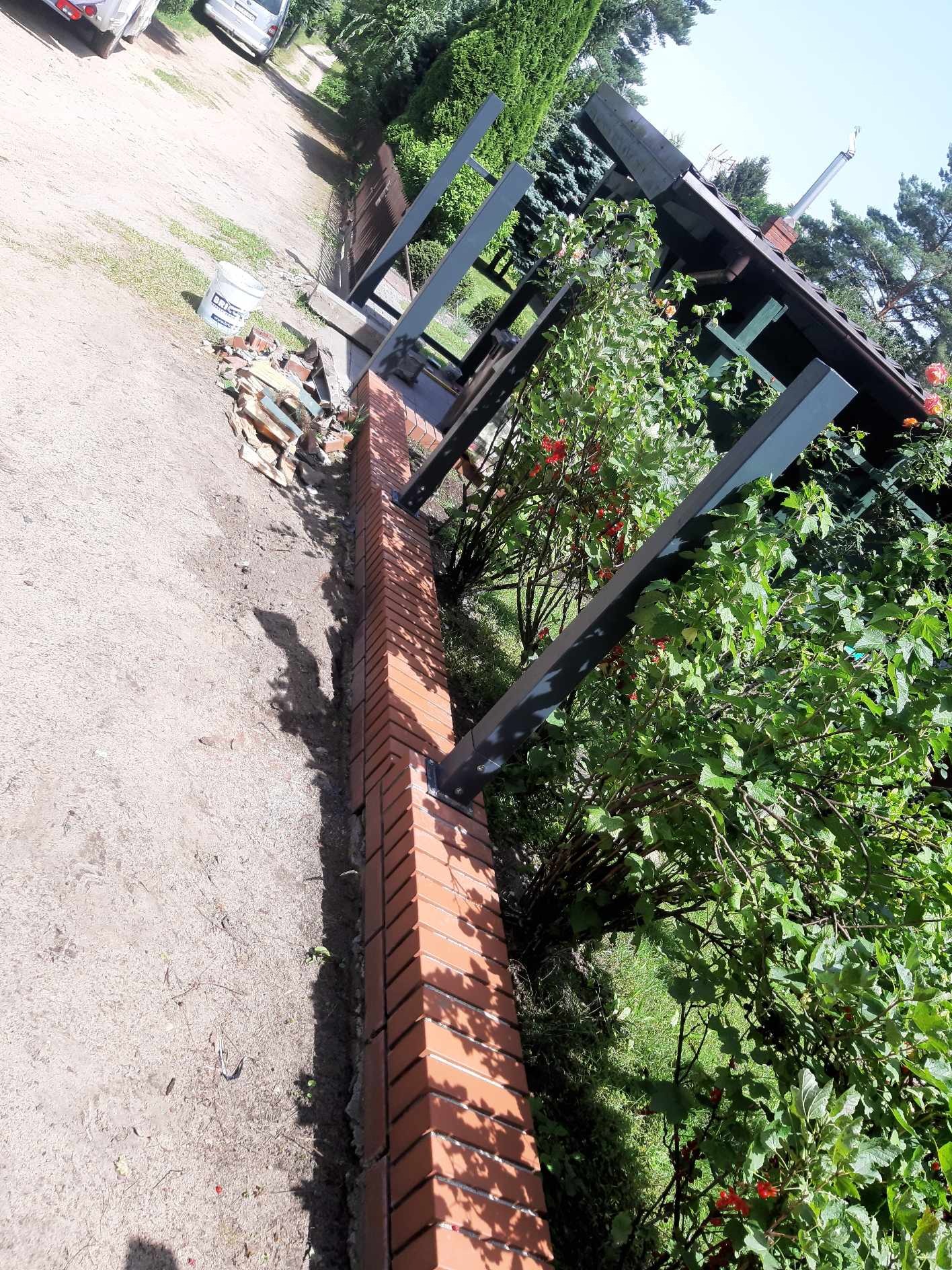 Wznoszenie murowanego ogrodzenia z cegły klinkierowej z metalowymi słupkami pod zadaszenie, widoczne materiały budowlane obok.