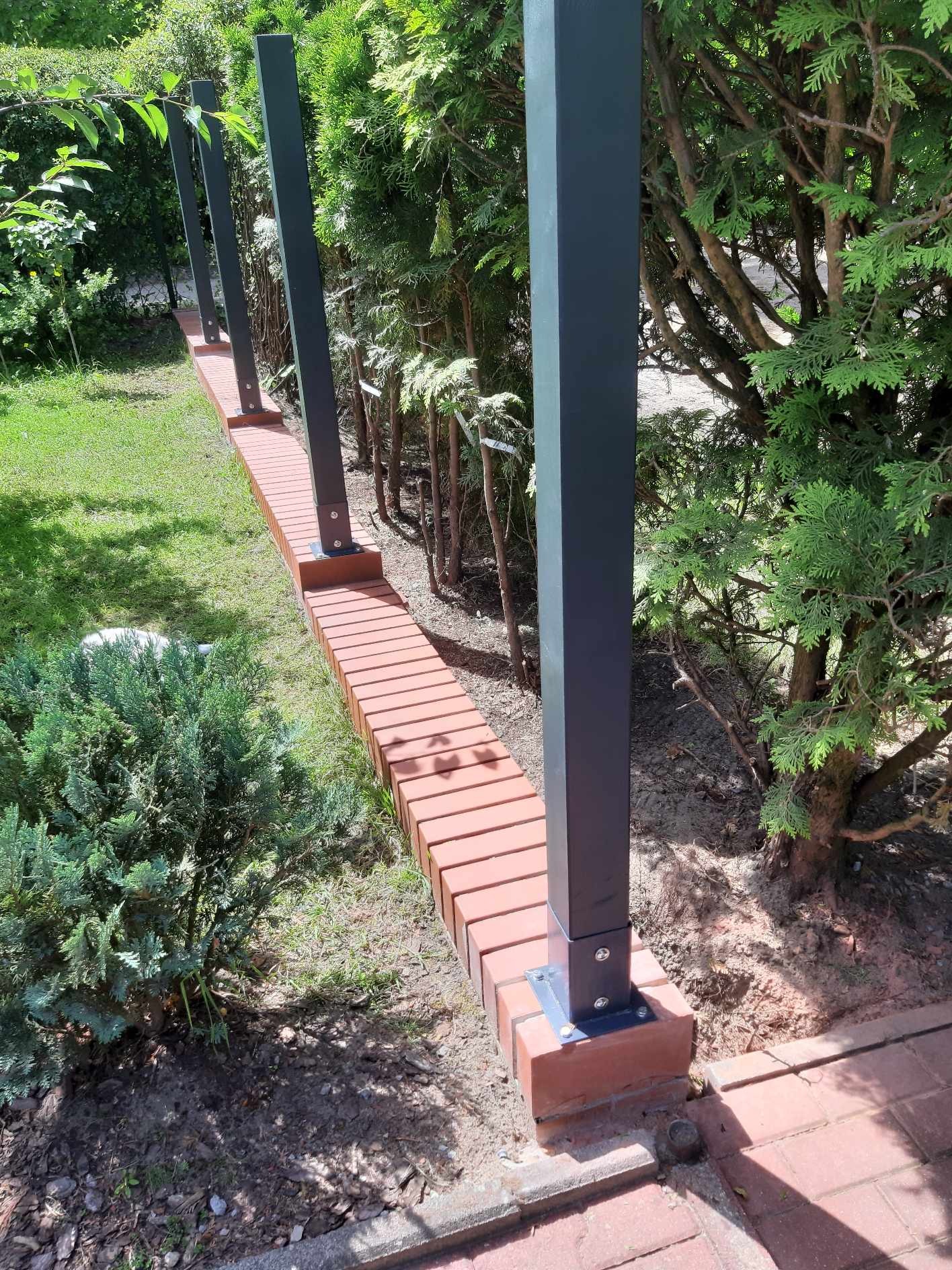 Nowoczesne ogrodzenie z cegły klinkierowej i metalowych słupków, otoczone zielenią w ogrodzie.