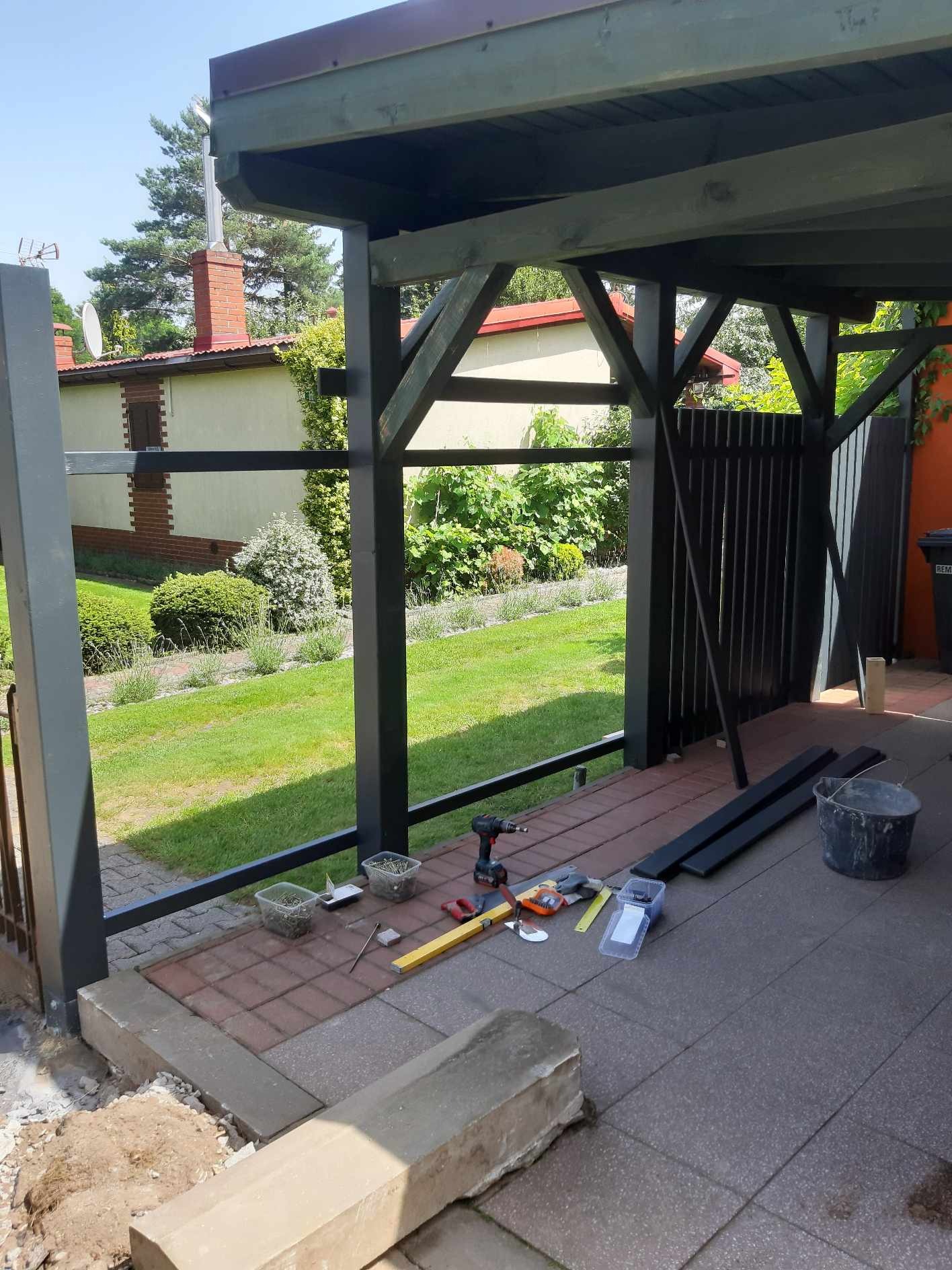 Montaż balustrady na tarasie: widoczne narzędzia, poziomica, wkrętarka, wiadro, elementy balustrady, kostka brukowa i trawnik w tle.