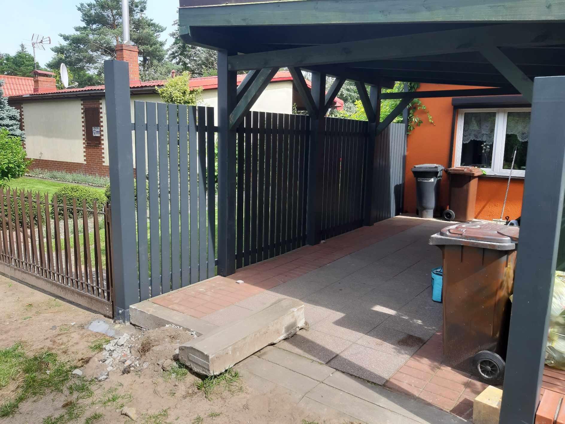 Nowoczesne ogrodzenie panelowe w kolorze grafitowym z bramą wjazdową, widoczne z zewnątrz posesji, z elementami kostki brukowej i betonowymi obrzeżami, fragment domu z elewacją w tle.