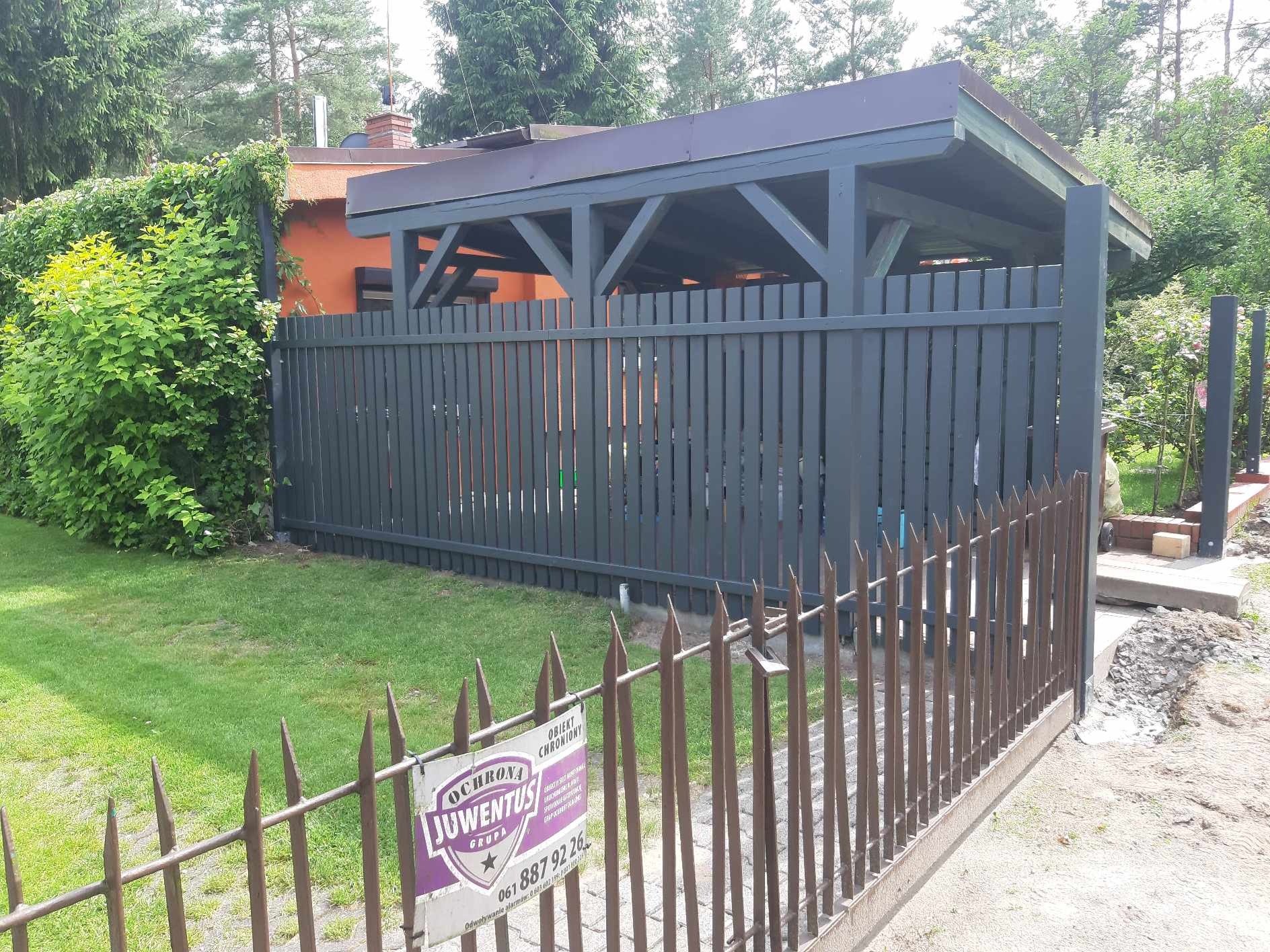 Grafitowa drewniana konstrukcja z zadaszeniem i ogrodzeniem panelowym, widoczna zza metalowego ogrodzenia z ostrymi zakończeniami w Poznaniu, z fragmentem pomarańczowego budynku w tle.