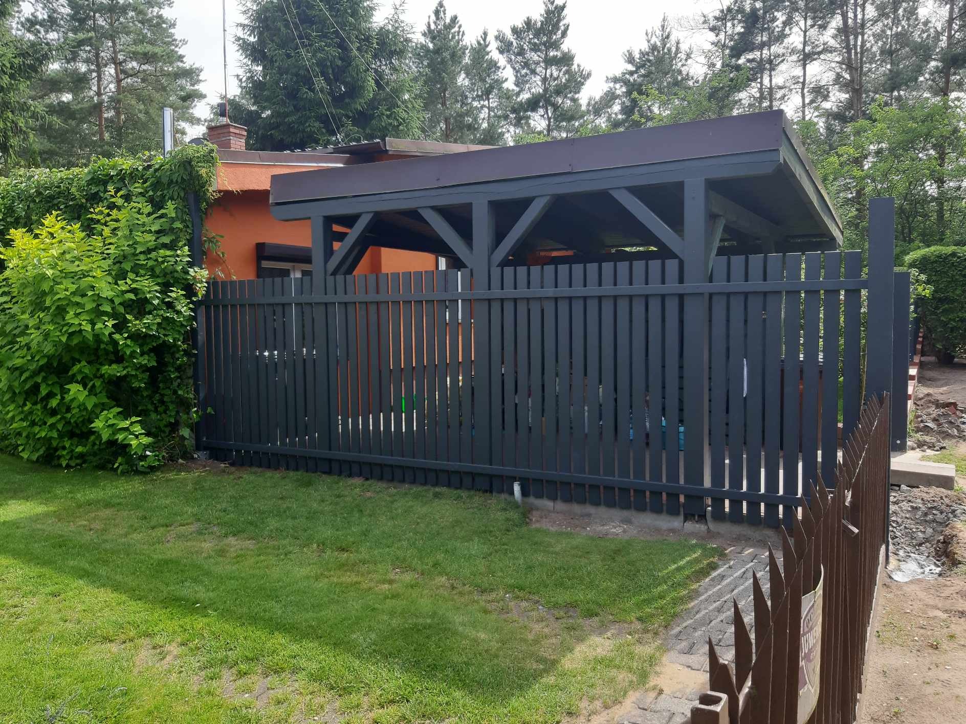 Ciemnoszary drewniany płot sztachetowy z zadaszeniem w tle, obok domu z pomarańczową elewacją i zielenią, widoczne ślady prac ziemnych.