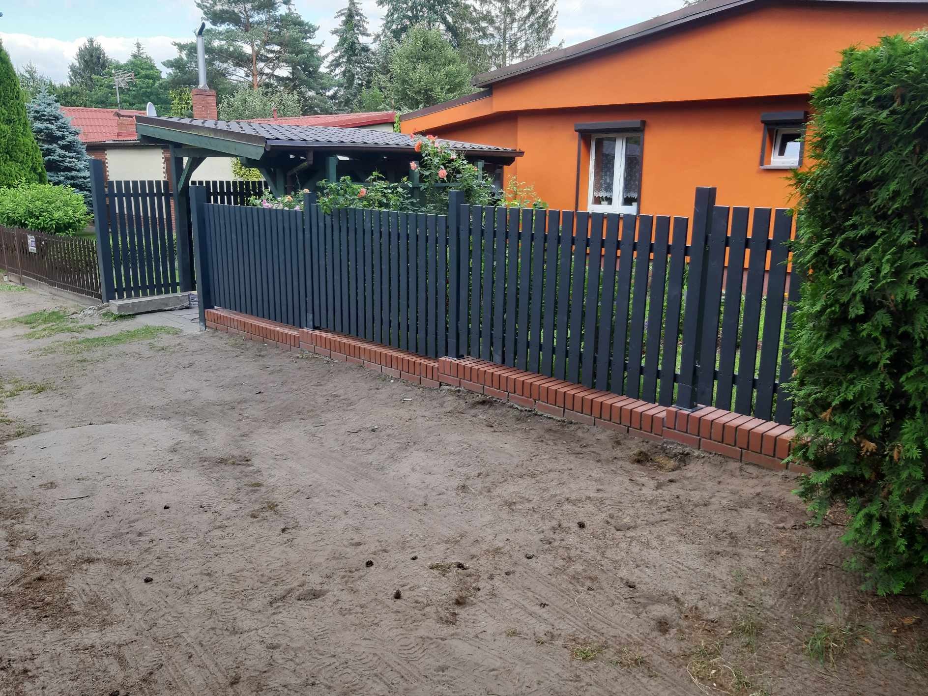 Nowo postawione, grafitowe ogrodzenie z ceglaną podmurówką na piaszczystym terenie, w tle pomarańczowy dom z białymi oknami i zielona roślinność.