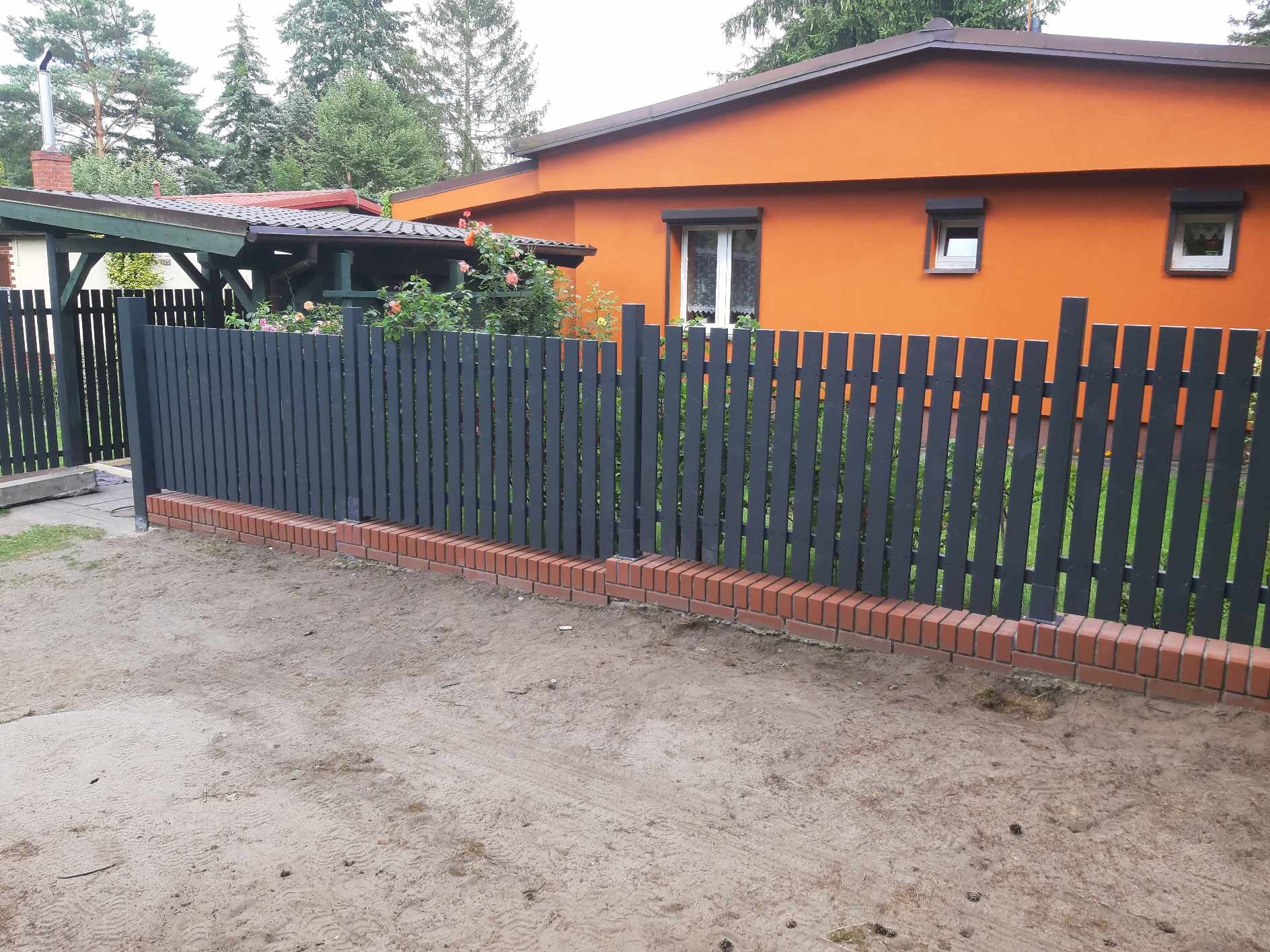 Ciemnoszary, drewniany płot sztachetowy na ceglanej podmurówce, oddzielający posesję z pomarańczowym domem, widok z poziomu gruntu.