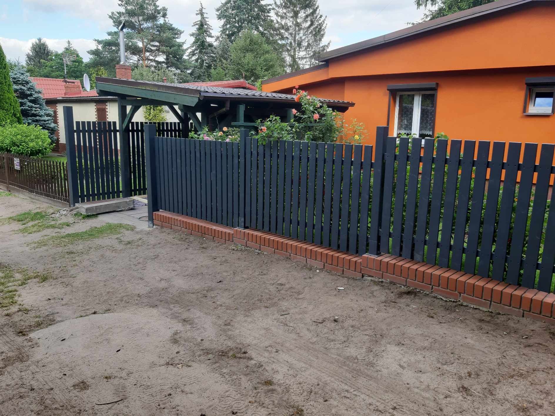 Ciemnoszary drewniany płot sztachetowy z ceglaną podmurówką na piaszczystym podłożu, w tle pomarańczowy dom i zieleń.