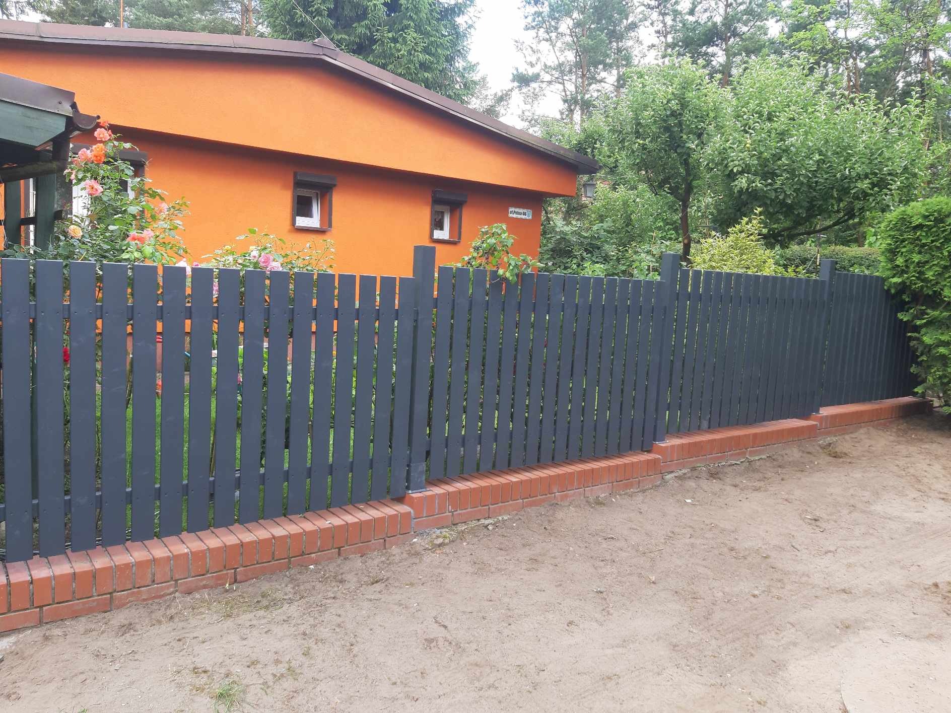 Szary, drewniany płot sztachetowy na ceglanej podmurówce, oddzielający posesję z pomarańczowym domem od piaszczystego terenu.