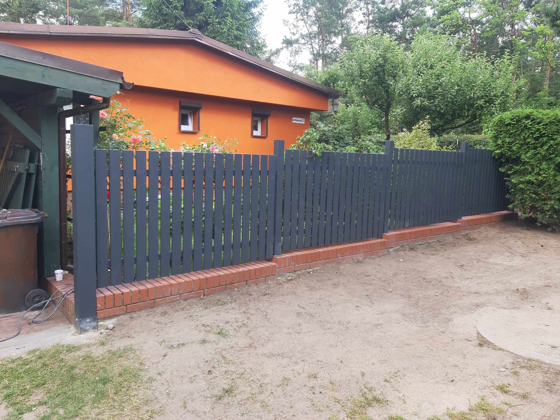 Nowoczesne, grafitowe ogrodzenie panelowe z podmurówką z czerwonej cegły, na tle pomarańczowego domu z kwitnącymi różami i zielenią drzew, widoczne ślady prac ziemnych.