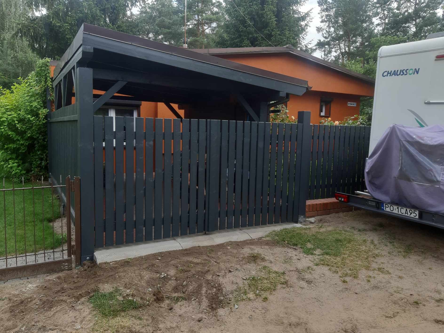 Nowoczesna, grafitowa brama posesji z drewnianych sztachet, zadaszona wiata garażowa i pomarańczowy budynek w tle, przyczepa kempingowa zaparkowana obok.