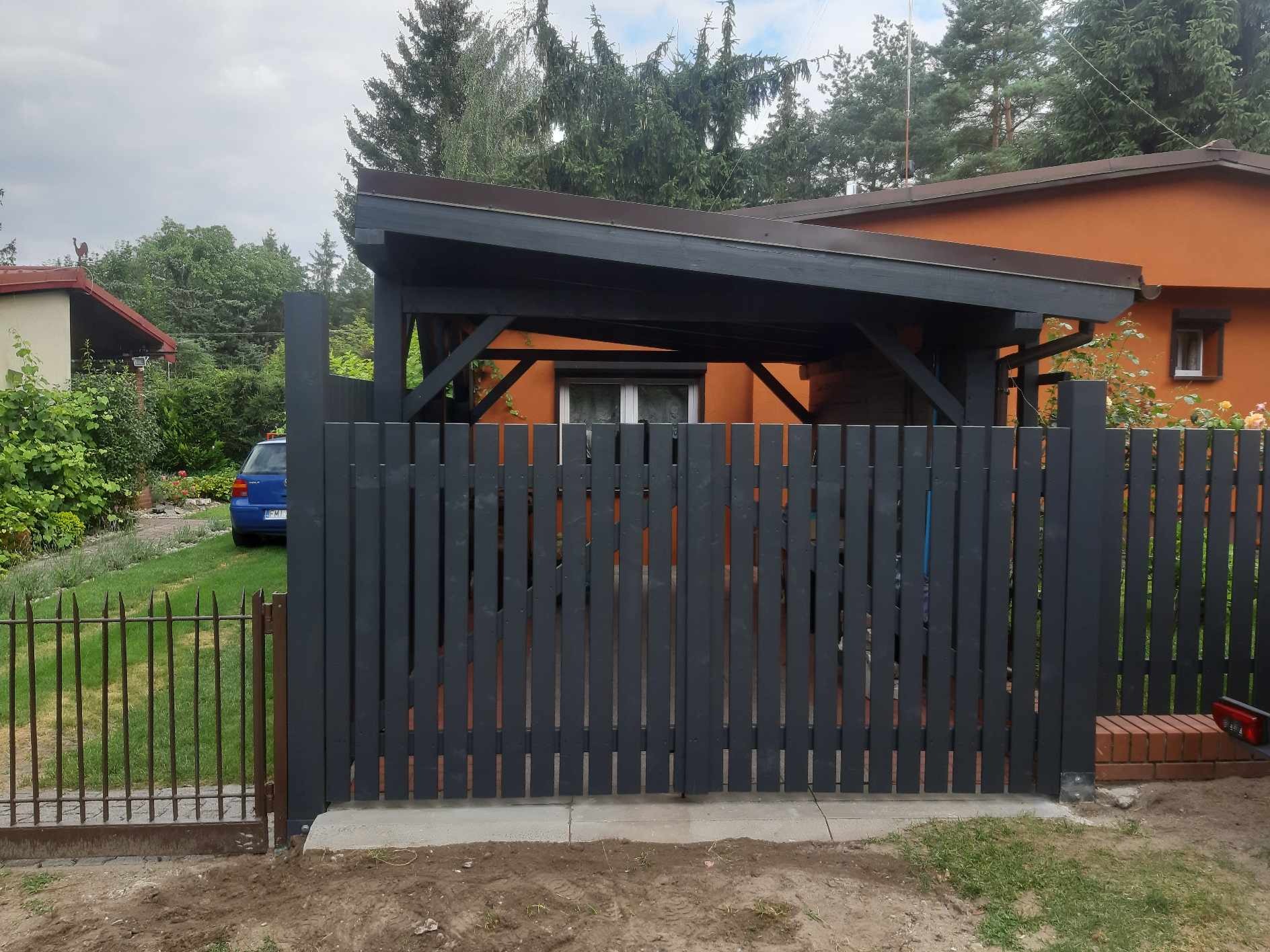 Ciemnoszara drewniana brama wjazdowa na posesję z widocznym fragmentem pomarańczowego domu i niebieskiego samochodu w tle.