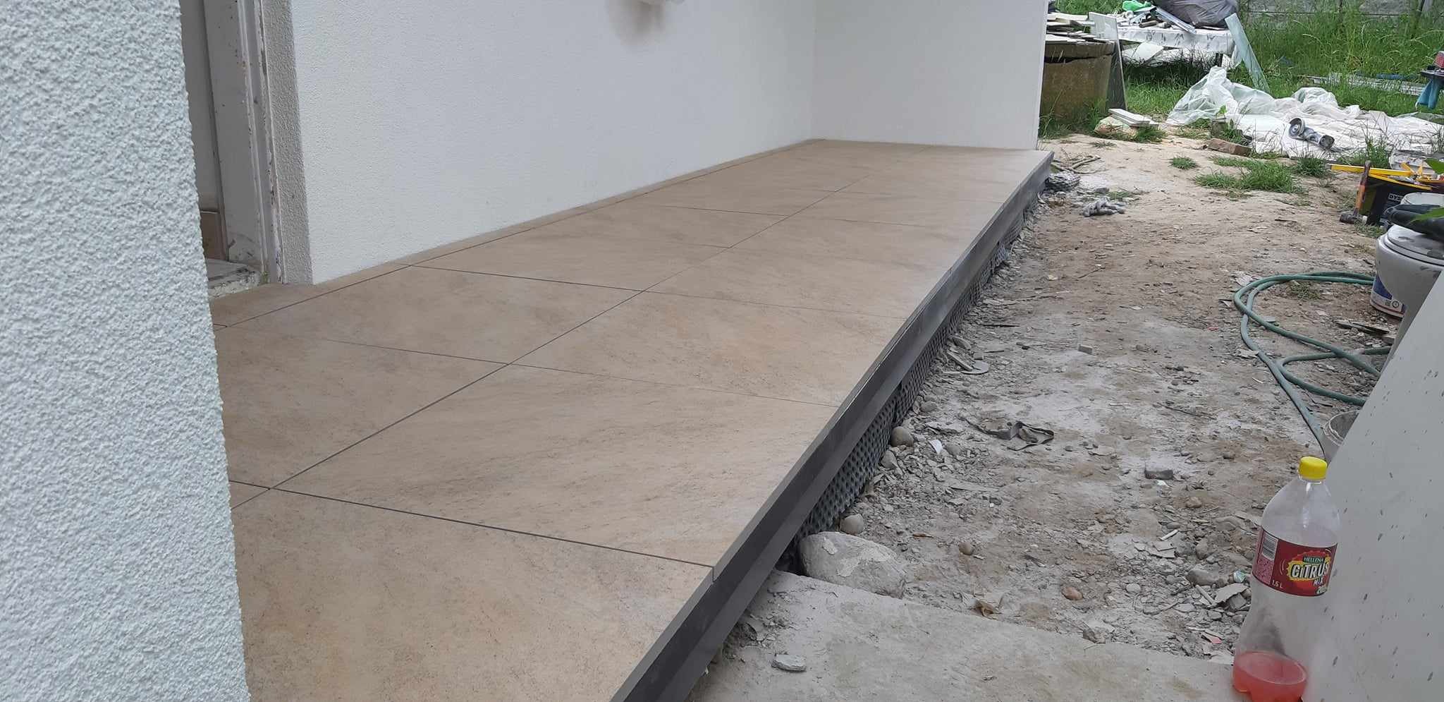 Wyłożony jasnymi płytkami taras przed budynkiem, widoczne detale wykończenia krawędzi i otoczenie w trakcie prac budowlanych.