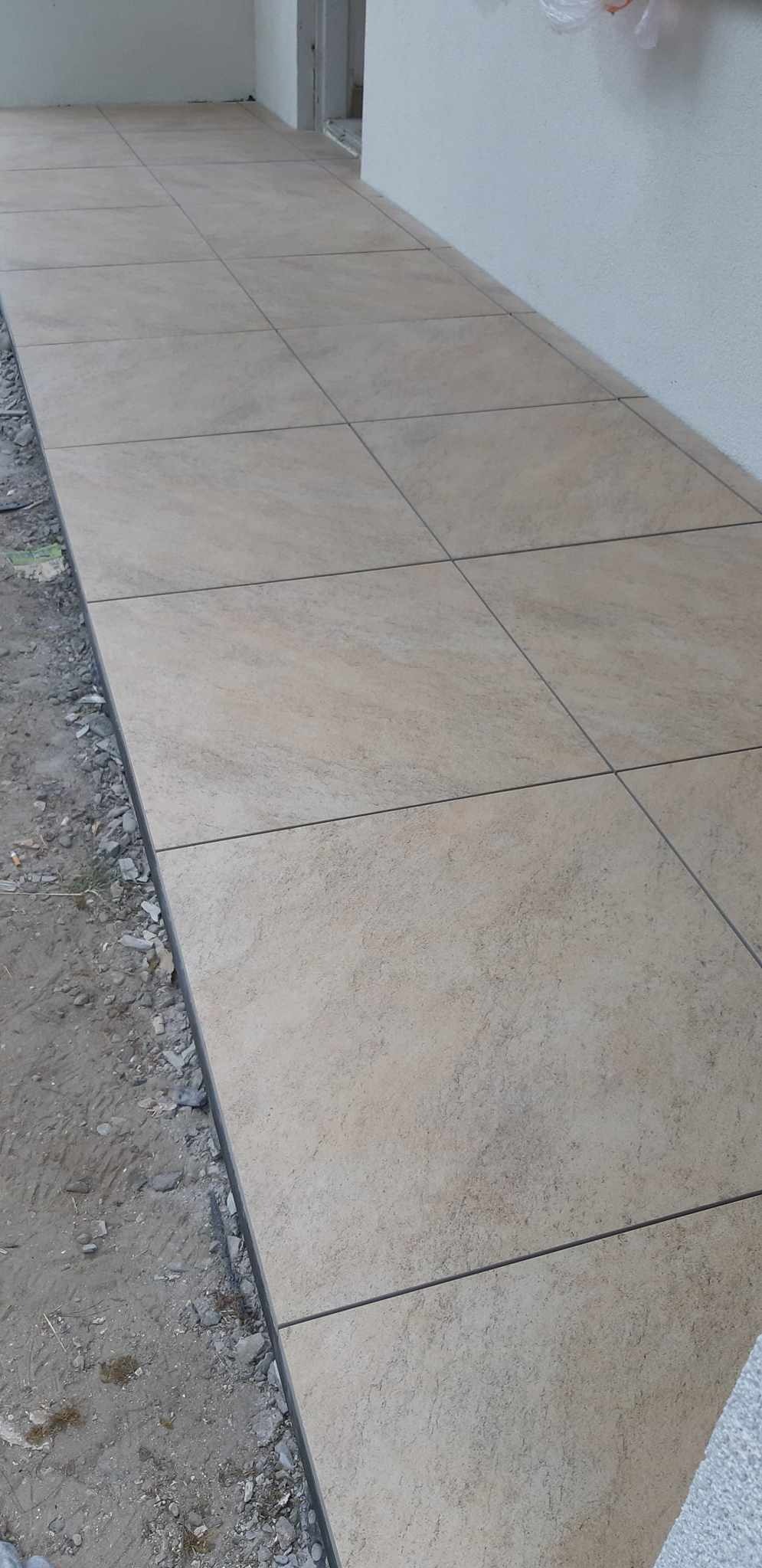 Ułożona podłoga z jasnych, kwadratowych płytek ceramicznych z ciemnymi fugami, widoczne niedokończone krawędzie i surowy grunt.