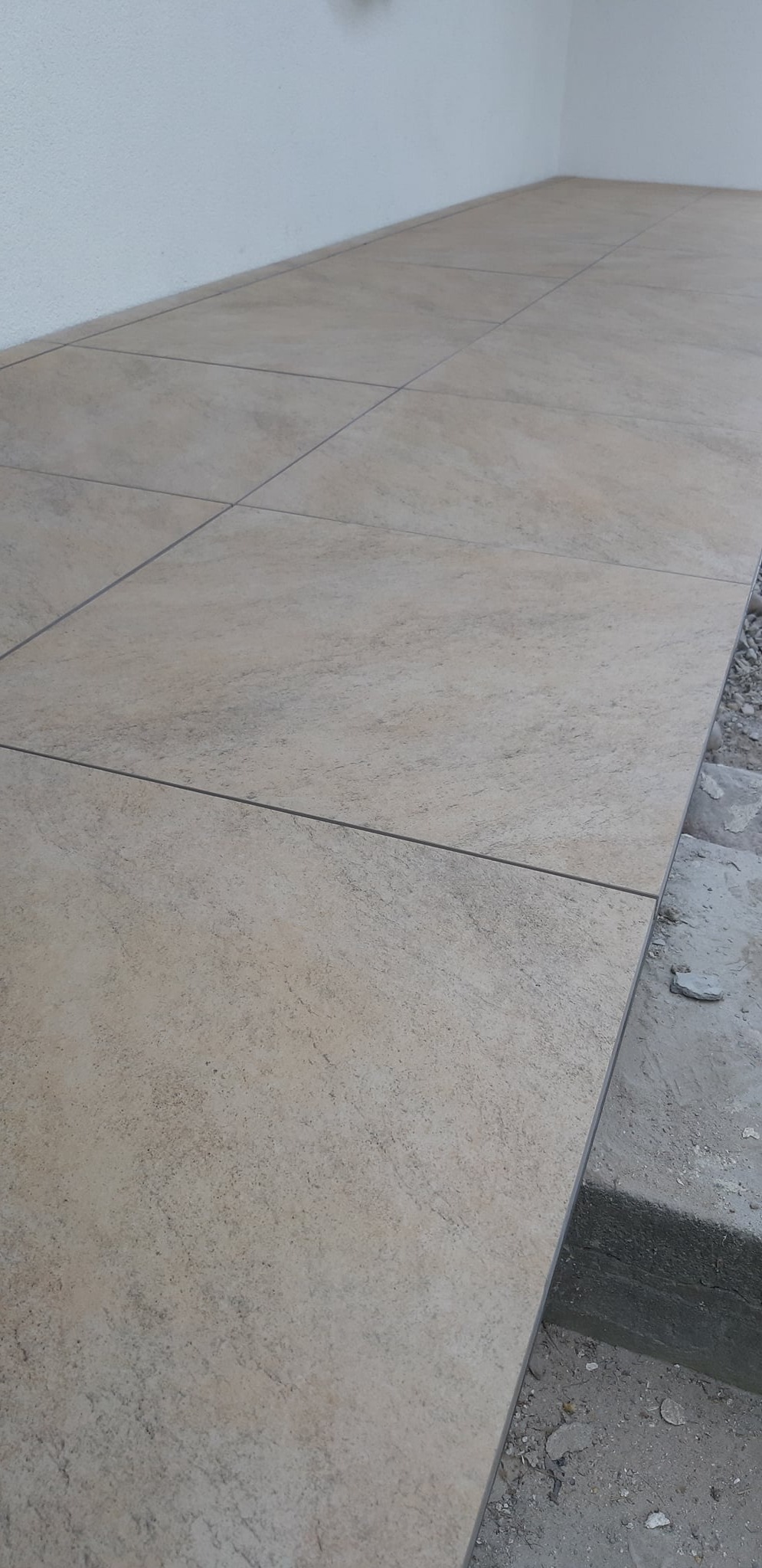 Beżowe płytki podłogowe o fakturze kamienia, ułożone w regularny wzór, widoczne szczeliny między płytkami, krawędź z surowym betonem.