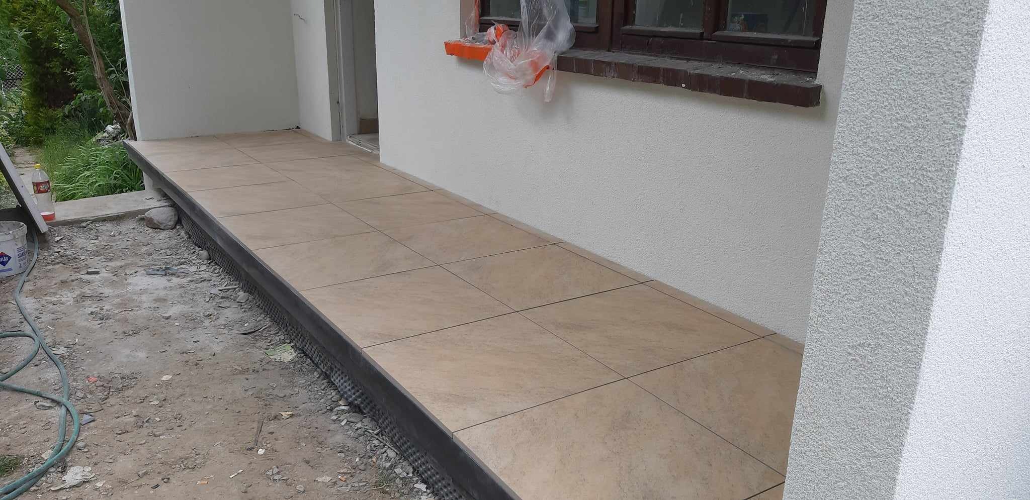 Wykończony taras z beżowych płytek ceramicznych, widoczne detale listwy krawędziowej i fragment elewacji z oknem zabezpieczonym folią malarską.