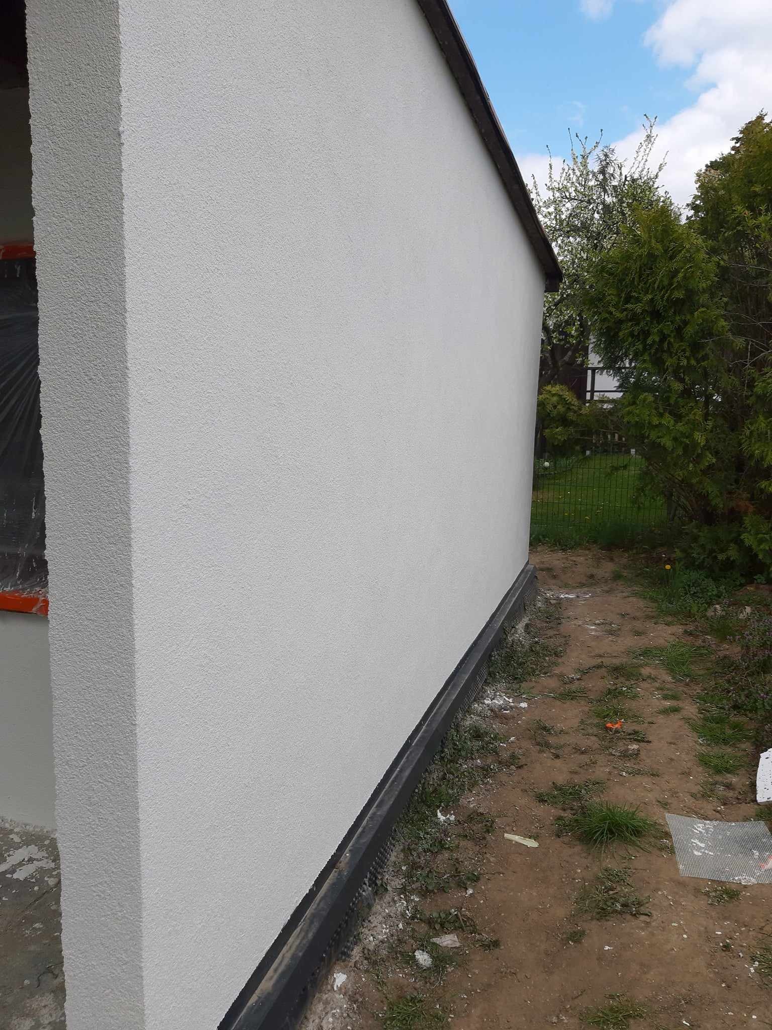 Świeżo otynkowana, jasnoszara ściana budynku z widoczną fakturą, czarną listwą przypodłogową i fragmentem okna zabezpieczonego folią malarską; w tle zieleń ogrodu i błękitne niebo.