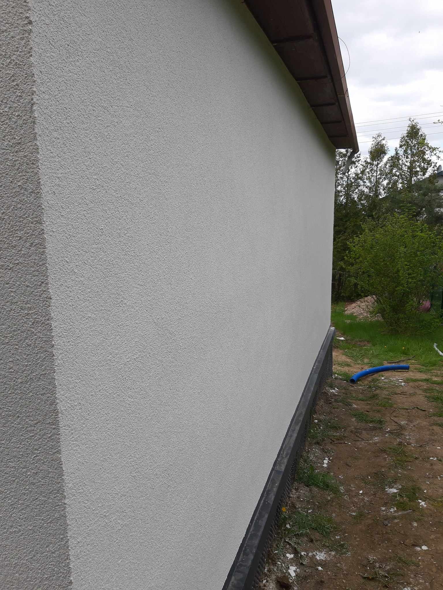 Świeżo wykonana, jasnoszara elewacja budynku jednorodzinnego z widocznym fragmentem dachu i otoczeniem z zielenią oraz błękitnym wężem spustowym na ziemi.