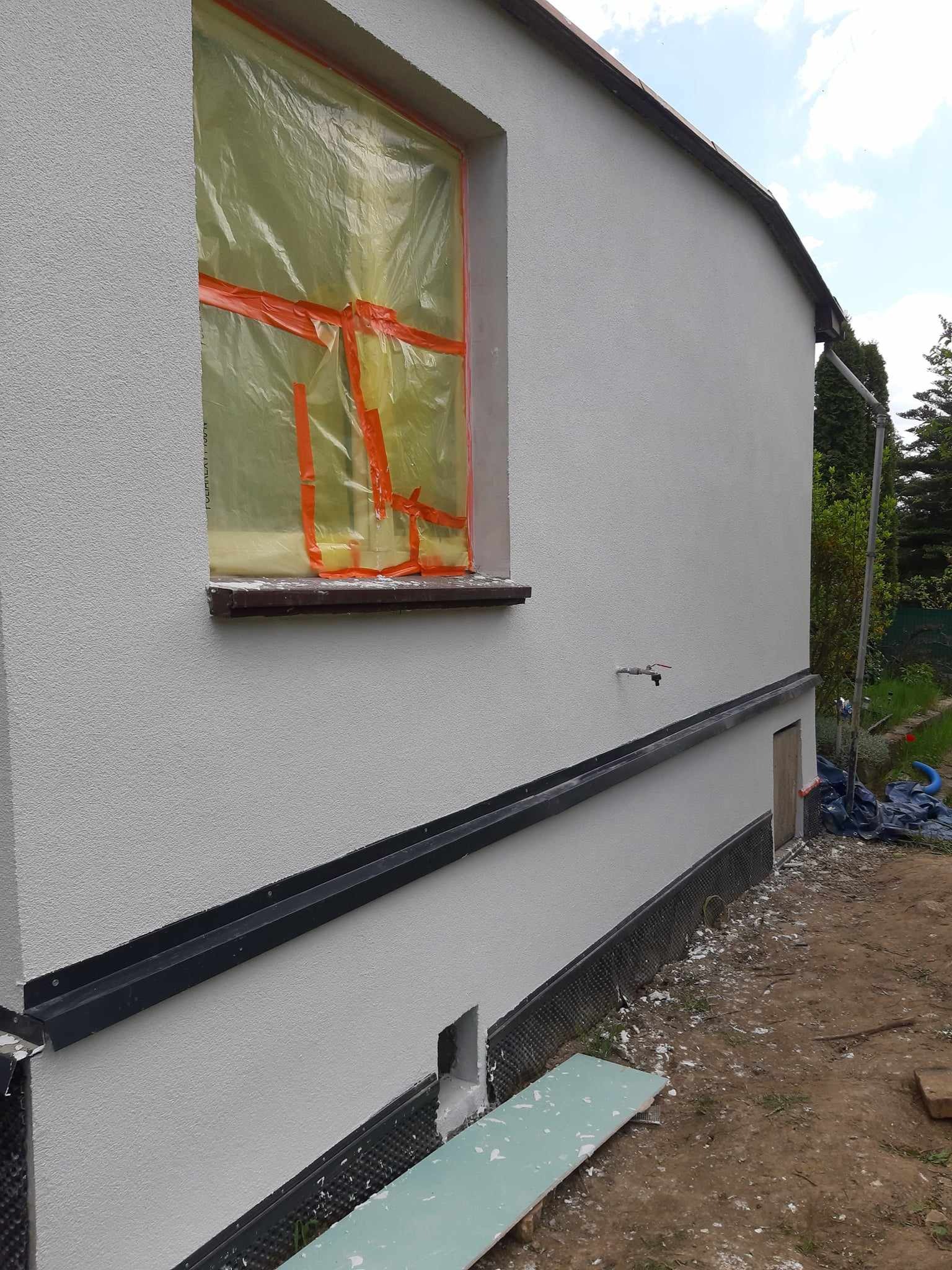 Ściana budynku po nałożeniu nowego tynku, okno zabezpieczone folią ochronną, widoczna listwa przypodłogowa i fragment terenu wokół budynku.