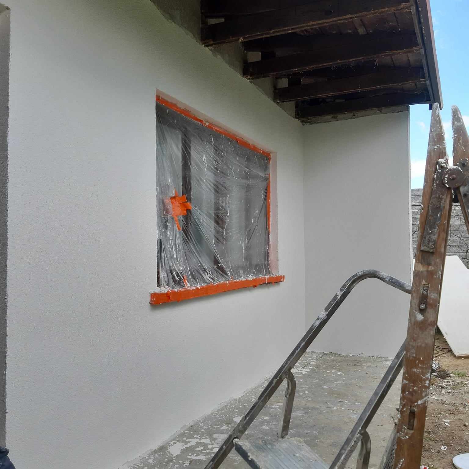 Świeżo otynkowana ściana budynku z zabezpieczonym oknem folią i pomarańczową taśmą, widoczne fragmenty drabiny i sufitu, jasne niebo w tle.