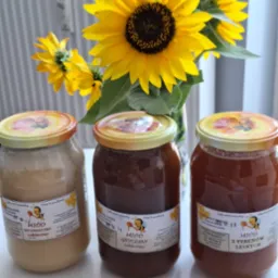 Trzy słoiki z różnymi rodzajami miodu (wielokwiatowy, gryczany, leśny) ustawione obok bukietu słoneczników w wazonie, na białym tle.