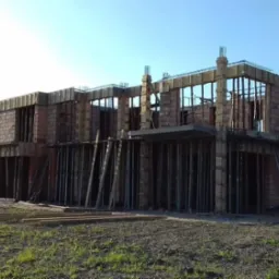 Dwukondygnacyjny budynek w trakcie budowy z czerwonej cegły, z widocznymi żelbetowymi słupami i deskowaniem, częściowo zadaszony, na tle zielonej trawy i błękitnego nieba.