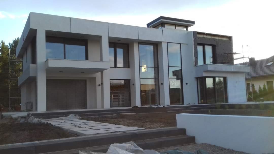 Nowoczesny dom z dużymi oknami i fragmentem elewacji w trakcie ocieplania, widoczne rusztowania i materiały budowlane.