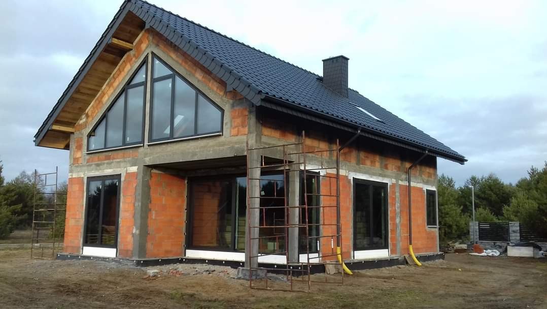 Niedokończony dom z cegły z czarnym dachem, widoczne rusztowanie i duże okna, izolacja fundamentów w trakcie realizacji.