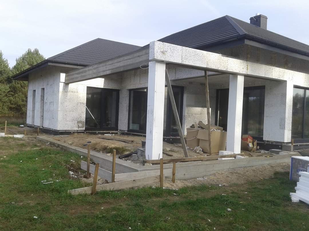 Wznoszenie ścian domu jednorodzinnego z betonu komórkowego, widoczne przygotowane fundamenty i częściowo zamontowane okna.