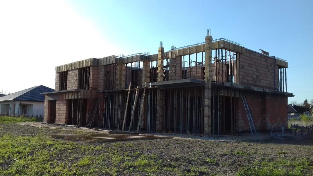 Dwukondygnacyjny budynek w trakcie budowy z czerwonej cegły, z widocznymi żelbetowymi słupami i deskowaniem, częściowo zadaszony, na tle zielonej trawy i błękitnego nieba.