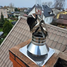 Mateusz Hopa - Nowy, błyszczący, metalowy komin z wiatrochronem na ceglanym kominie, na tle dachówki i zabudowy miejskiej w pochmurny dzień. Widok z góry.