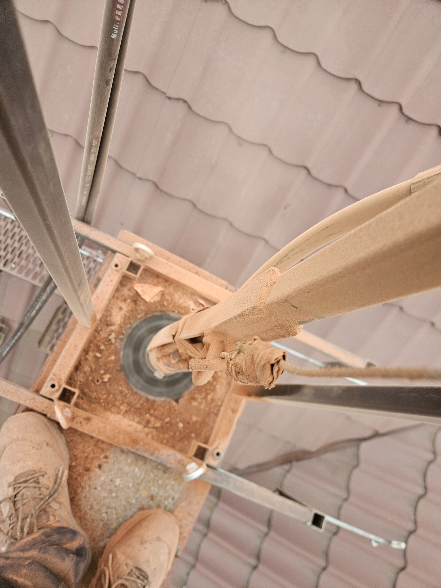 Widok z góry na komin na dachu z blachodachówki, z fragmentem rusztowania i butami pracownika, wszystko pokryte pyłem. Praca na wysokościach.