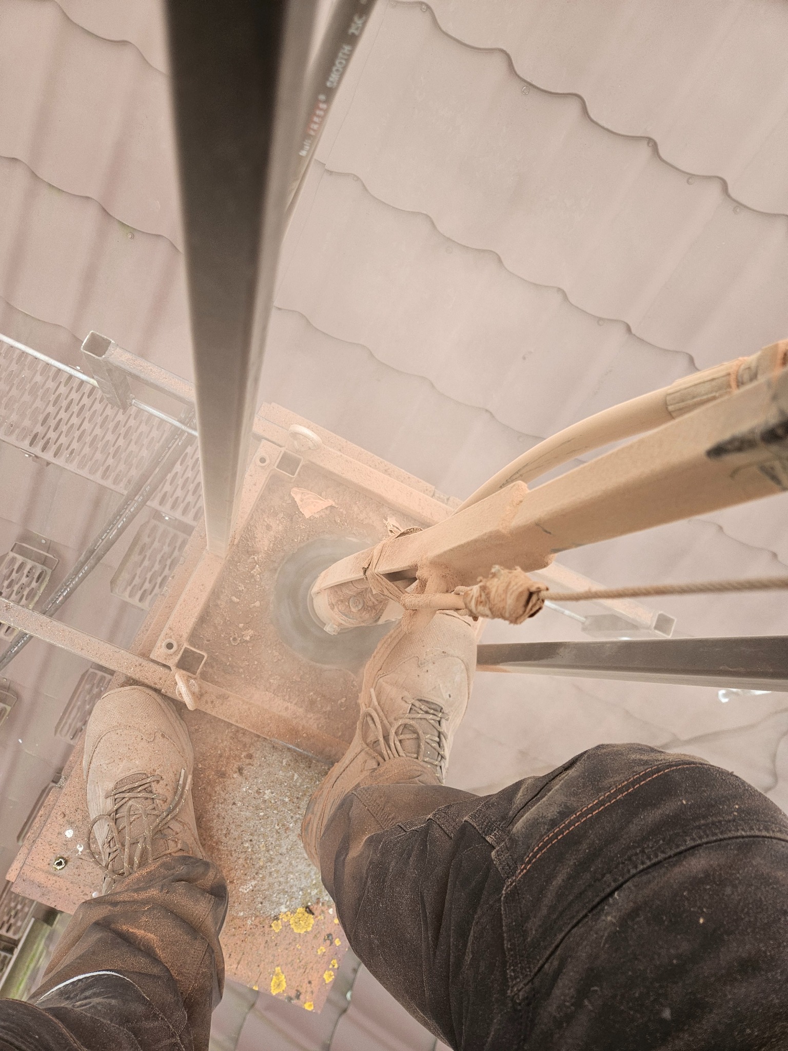 Widok z góry na kominiarza stojącego na dachu podczas pracy. Widoczne buty robocze, fragment drabiny i dach pokryty pyłem.