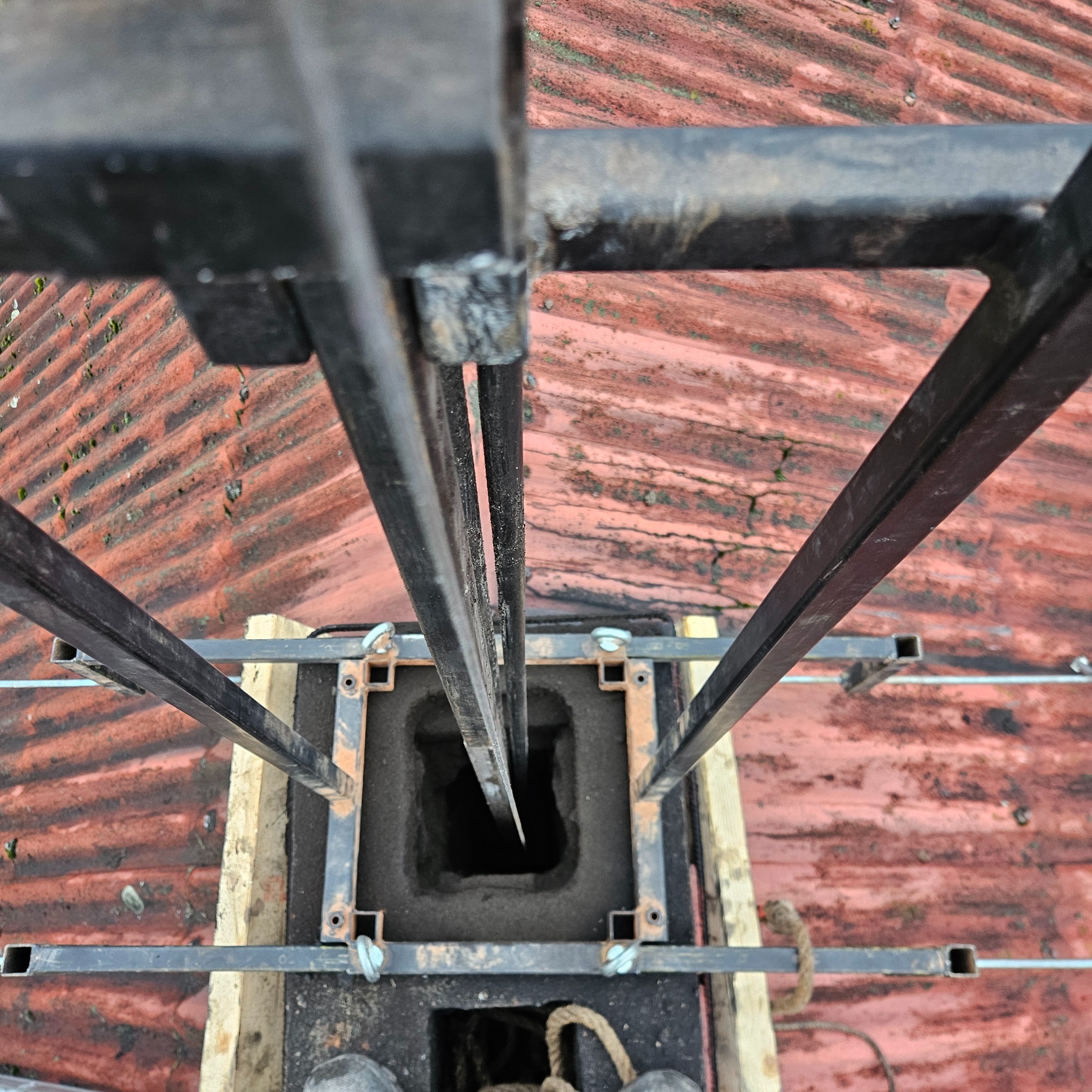 Widok z góry na komin z zamontowaną konstrukcją wsporczą na dachu z czerwonej dachówki. Widoczny otwór kominowy i elementy mocujące.