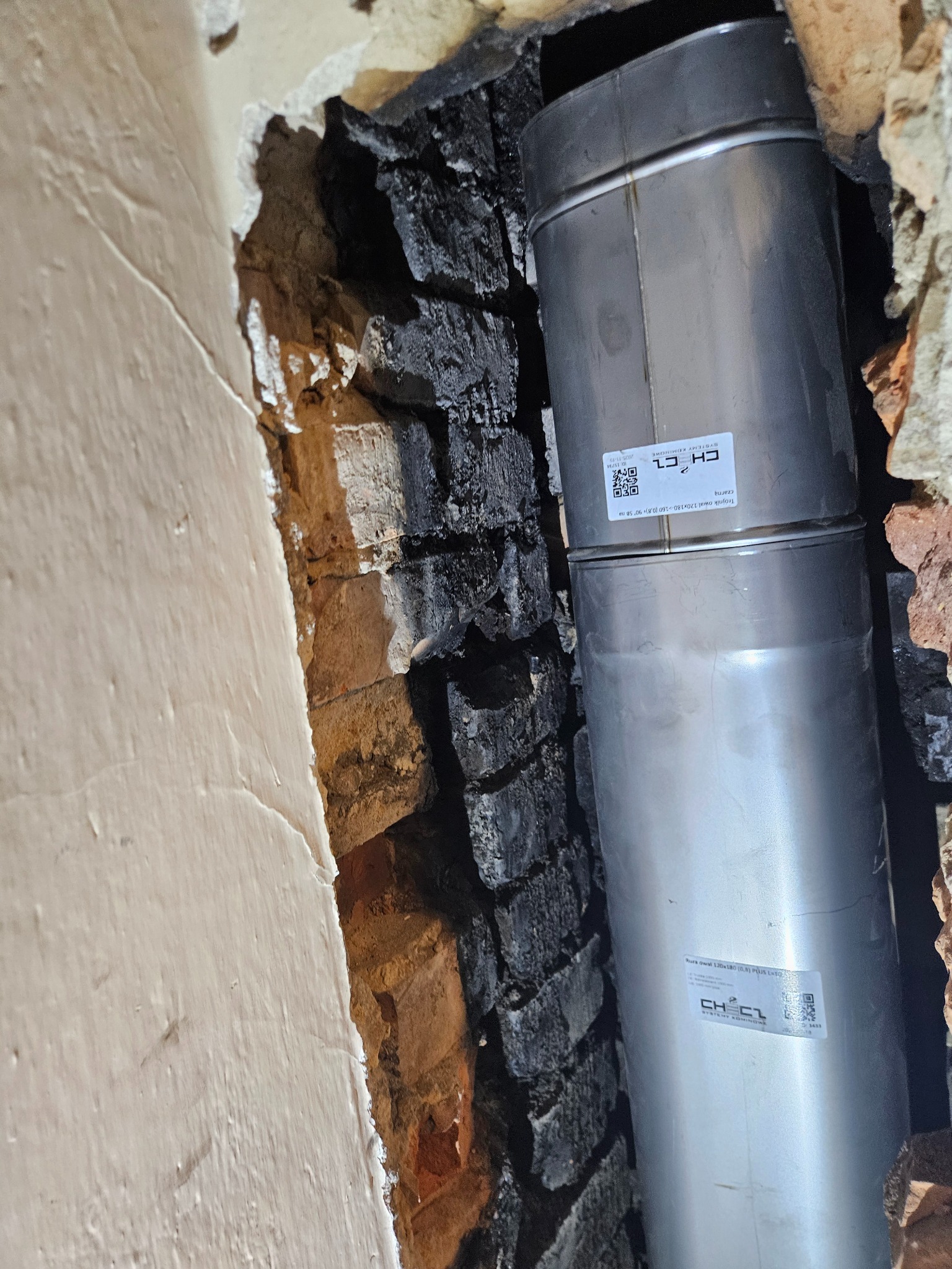 Wnętrze komina z nowym, metalowym wkładem kominowym. Widoczne ślady sadzy na cegłach i fragment otynkowanej ściany. Wkład z naklejką informacyjną.