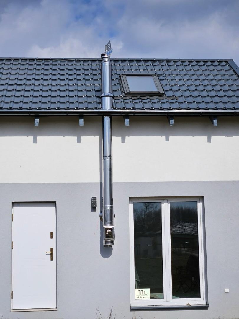 Zewnętrzny, stalowy komin systemowy zamontowany na elewacji domu z szarą dachówką. Widoczne okno dachowe i białe drzwi wejściowe. Słoneczny dzień.