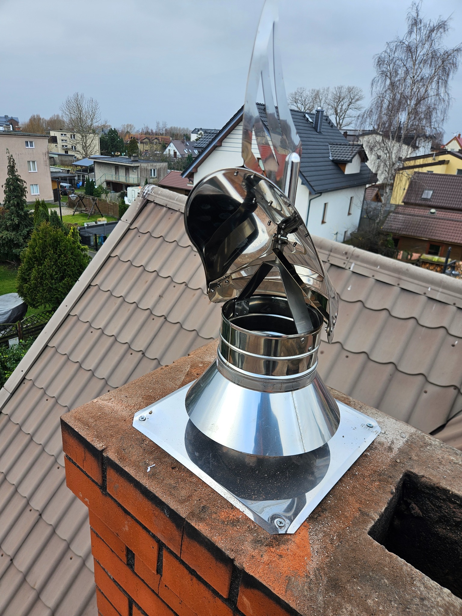 Nowy, błyszczący, metalowy komin z wiatrochronem na ceglanym kominie, na tle dachówki i zabudowy miejskiej w pochmurny dzień. Widok z góry.