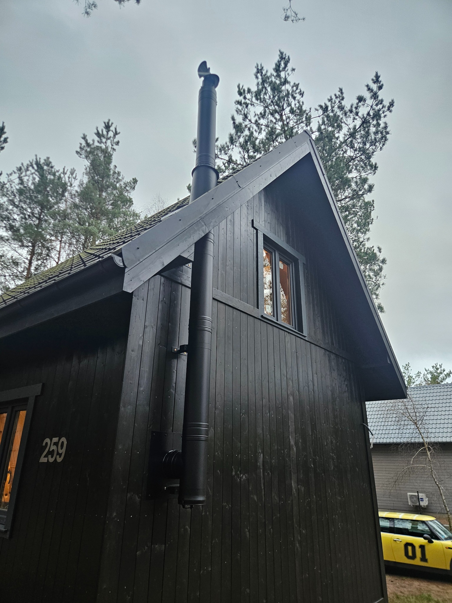 Ciemny, drewniany dom z czarnym kominem na zewnątrz, numer 259 na ścianie, widok z dołu, pochmurne niebo w tle. Komin zamontowany na elewacji.