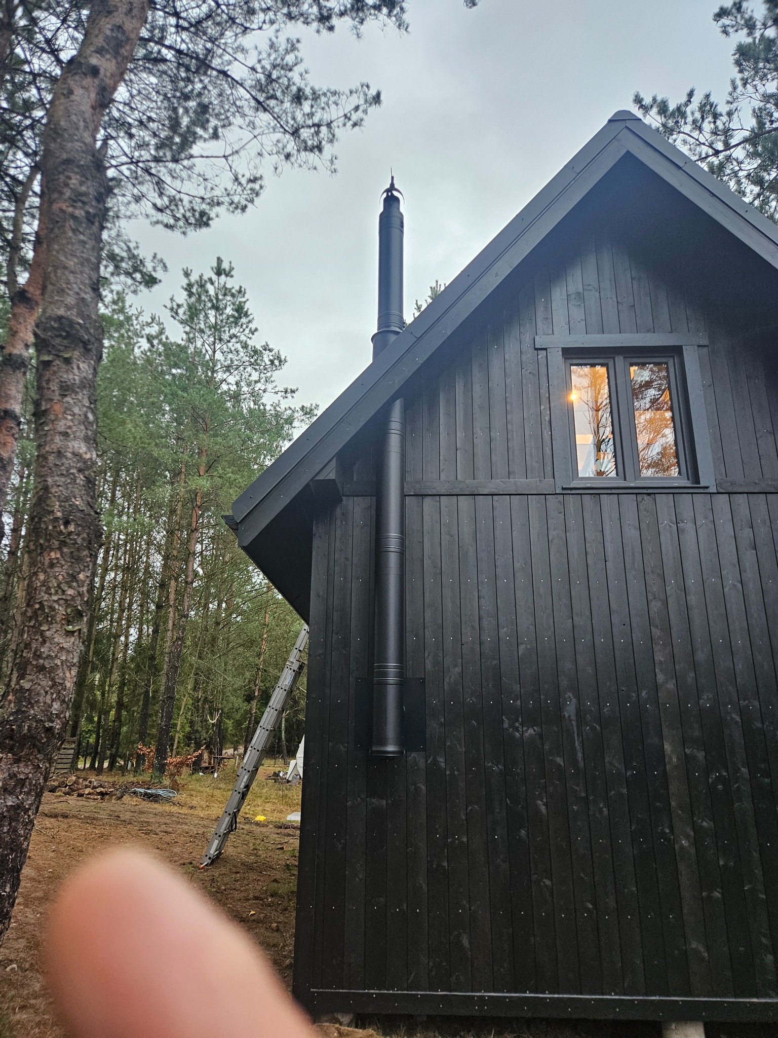 Ciemna, drewniana elewacja domu z kominem na tle lasu. W oknie odbija się ciepłe światło. Widoczna drabina oparta o ścianę. Ujęcie z dołu, z fragmentem palca w kadrze.