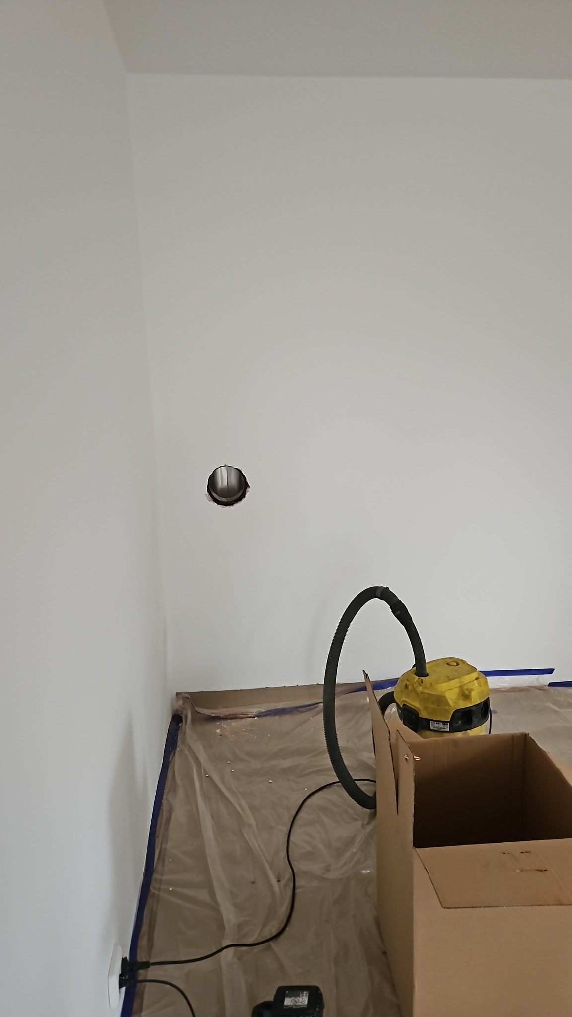 Otwór wentylacyjny w ścianie, zabezpieczona folią podłoga, odkurzacz i karton. Przygotowania do prac remontowych w pomieszczeniu.