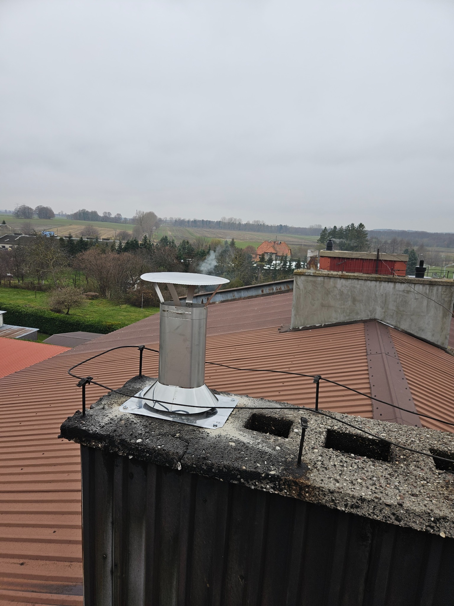 Błyszczący, metalowy komin z daszkiem na dachu z blachy trapezowej. W tle zabudowania i pola pod pochmurnym niebem. Widoczny dym wydobywający się z komina.