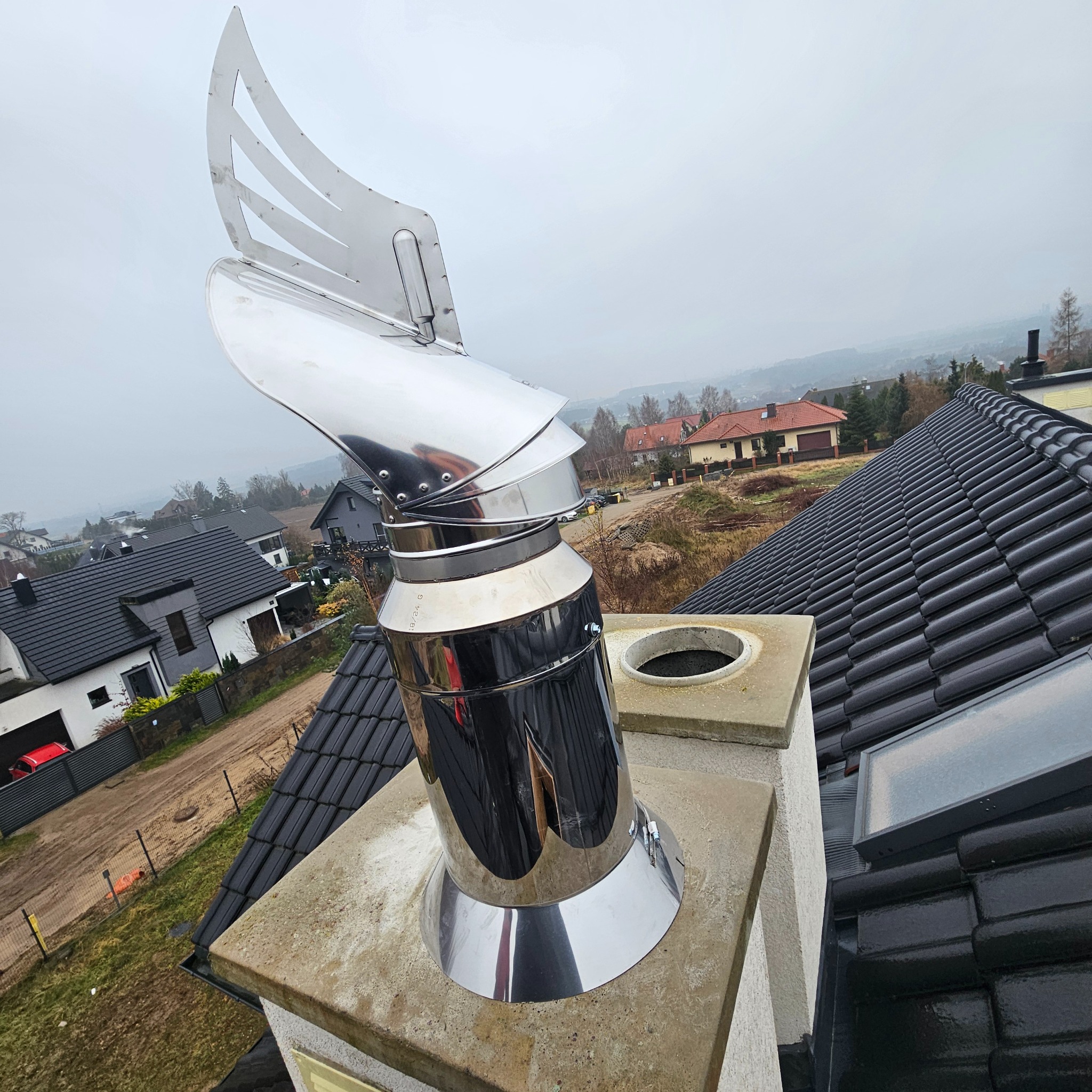 Błyszczący, metalowy komin z ozdobnym wiatrowskazem na dachu z czarnej dachówki, na tle zabudowań i pochmurnego nieba. Widok z góry.
