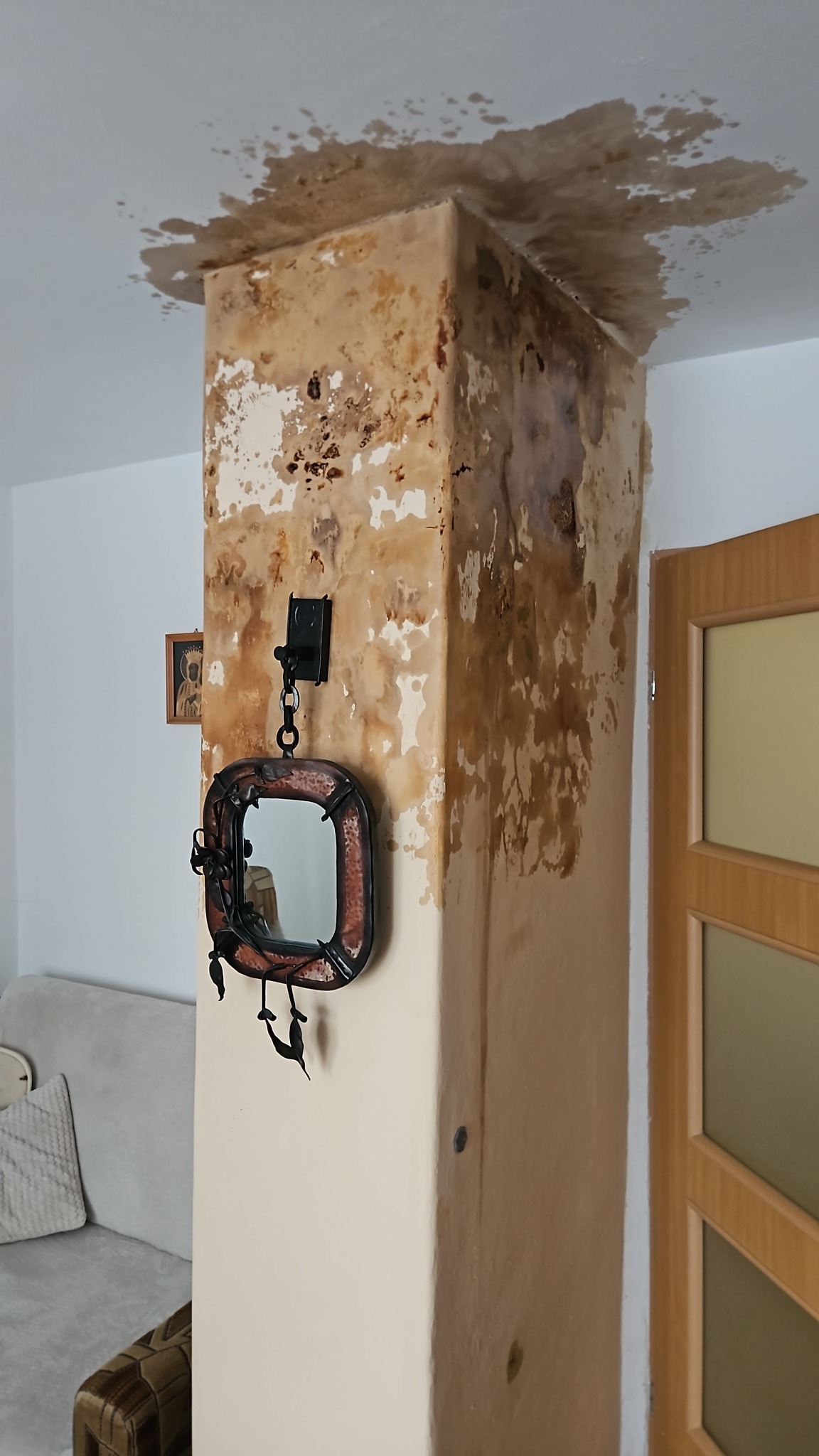 Zacieki na suficie i ścianie kolumny w pokoju. Widoczne plamy i odpadający tynk. Na ścianie wisi ozdobne lustro. Uszkodzenia po zalaniu.