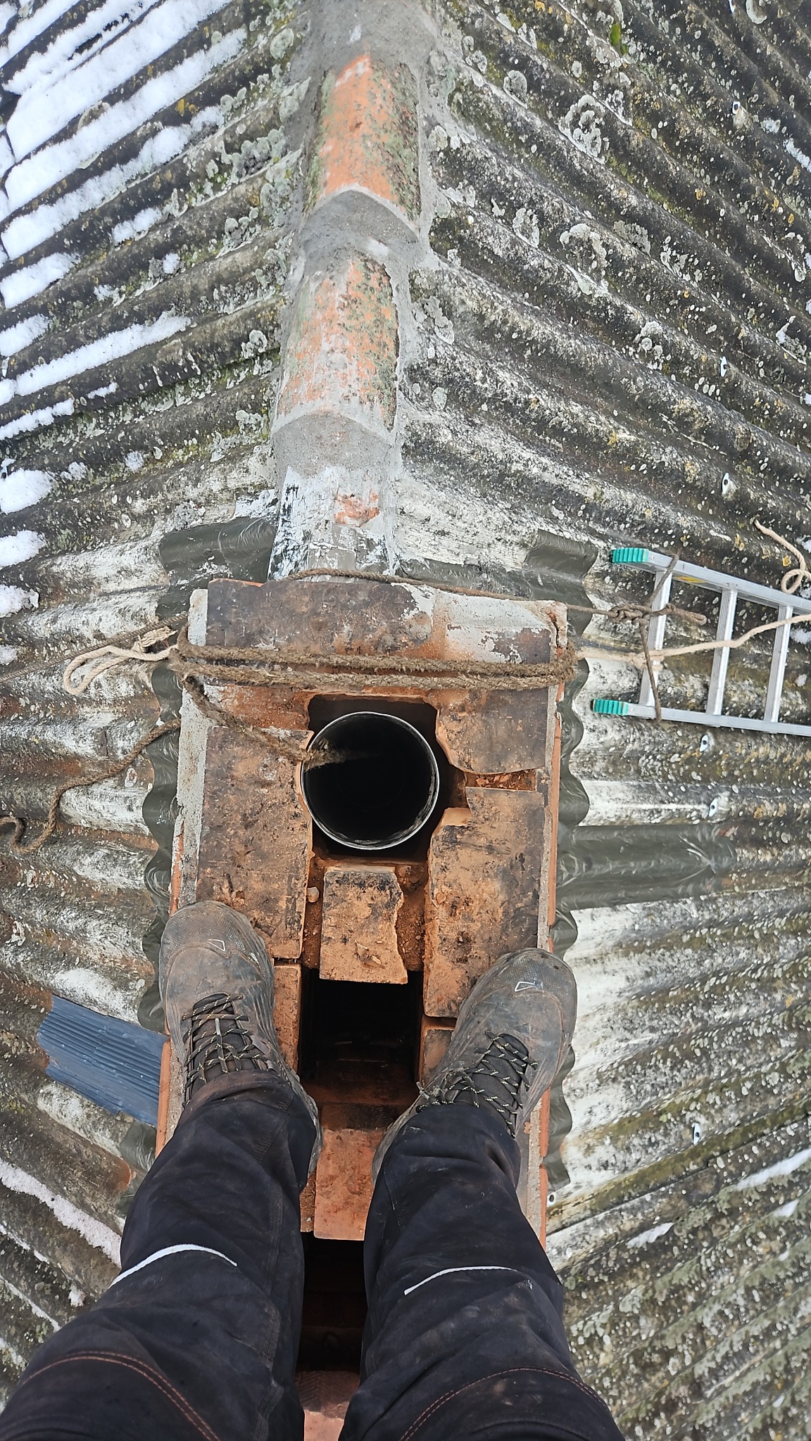 Widok z góry na komin z wkładem, buty na dachu z eternitu, lina zabezpieczająca i drabina. Praca na wysokości, widoczne zabrudzenia i ślady użytkowania.