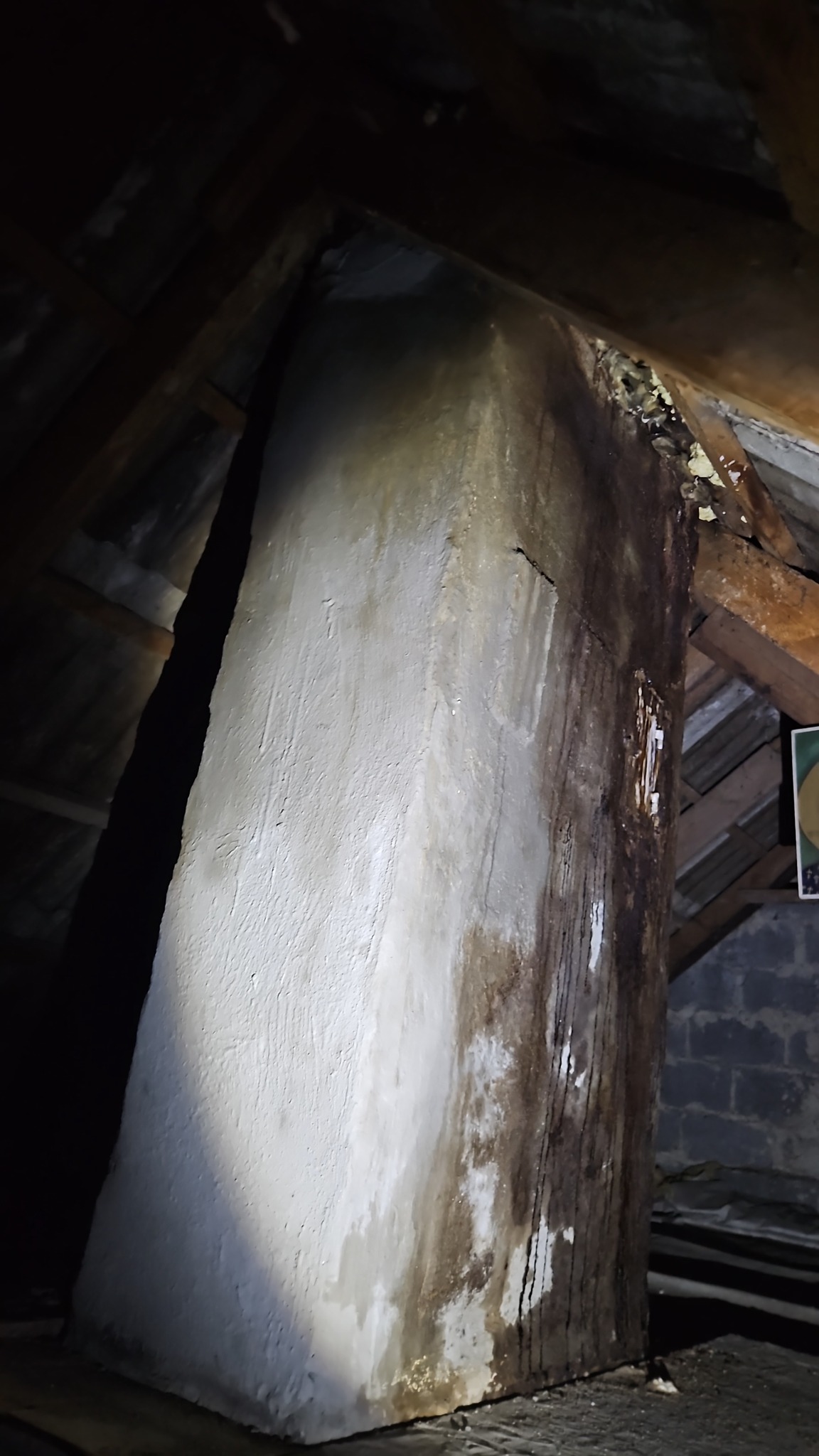 Oczyszczony komin w Lęborku na poddaszu, z widocznymi śladami po zalaniu. Komin otynkowany na biało, z ciemnymi zaciekami. Elementy drewnianej konstrukcji dachu.
